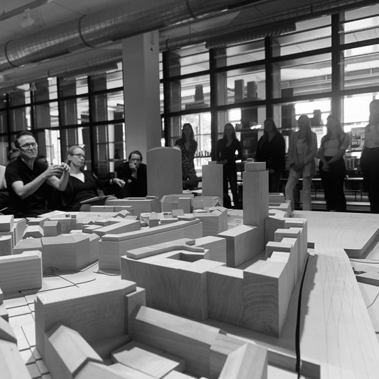Im Vordergrund des Bildes ist ein Stadtmodell aus Holz zu sehen, über das ein Teil des Teams mit Studierenden im Hintergrund diskutiert.